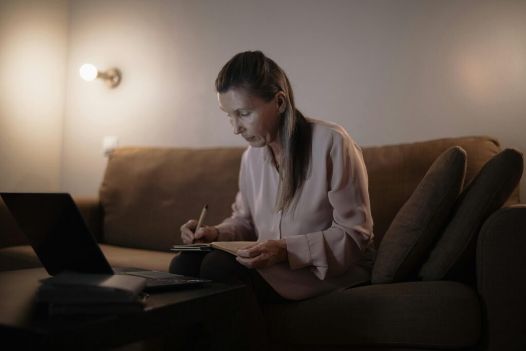 Senior woman sitting on couch, writing notes while using a laptop, cozy indoor setting.