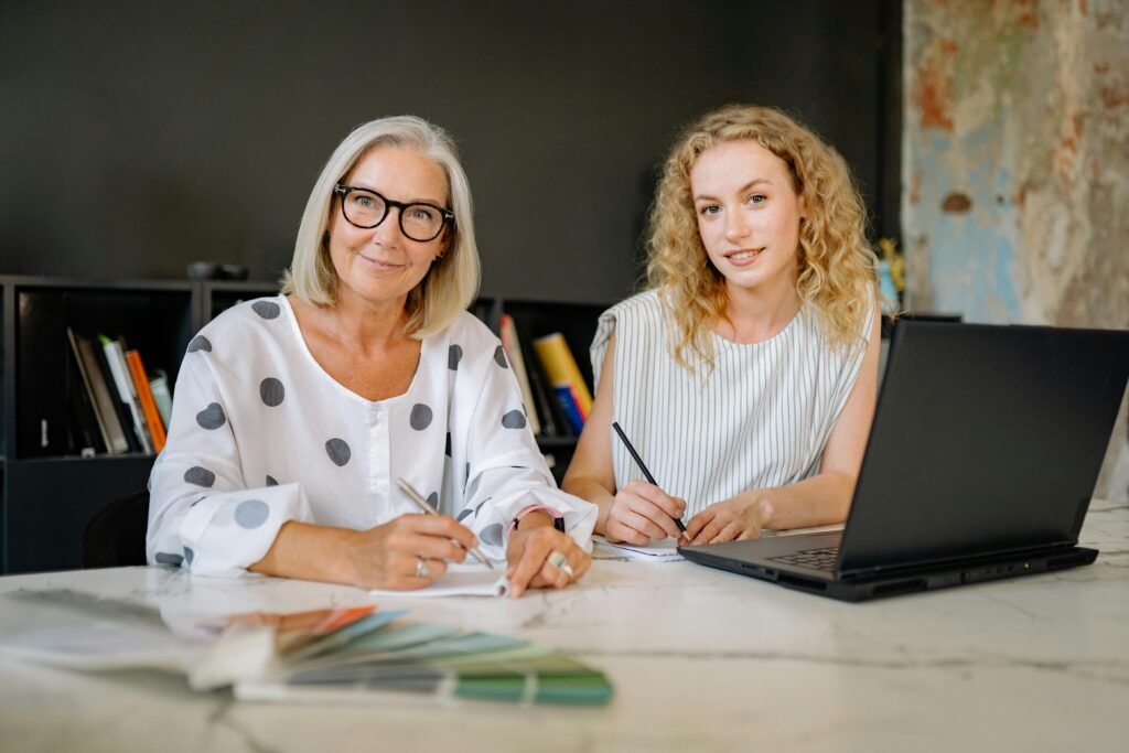 pexels-photo-8837490-8837490 Two women smiling and working together in a modern office setting with a laptop and documents.